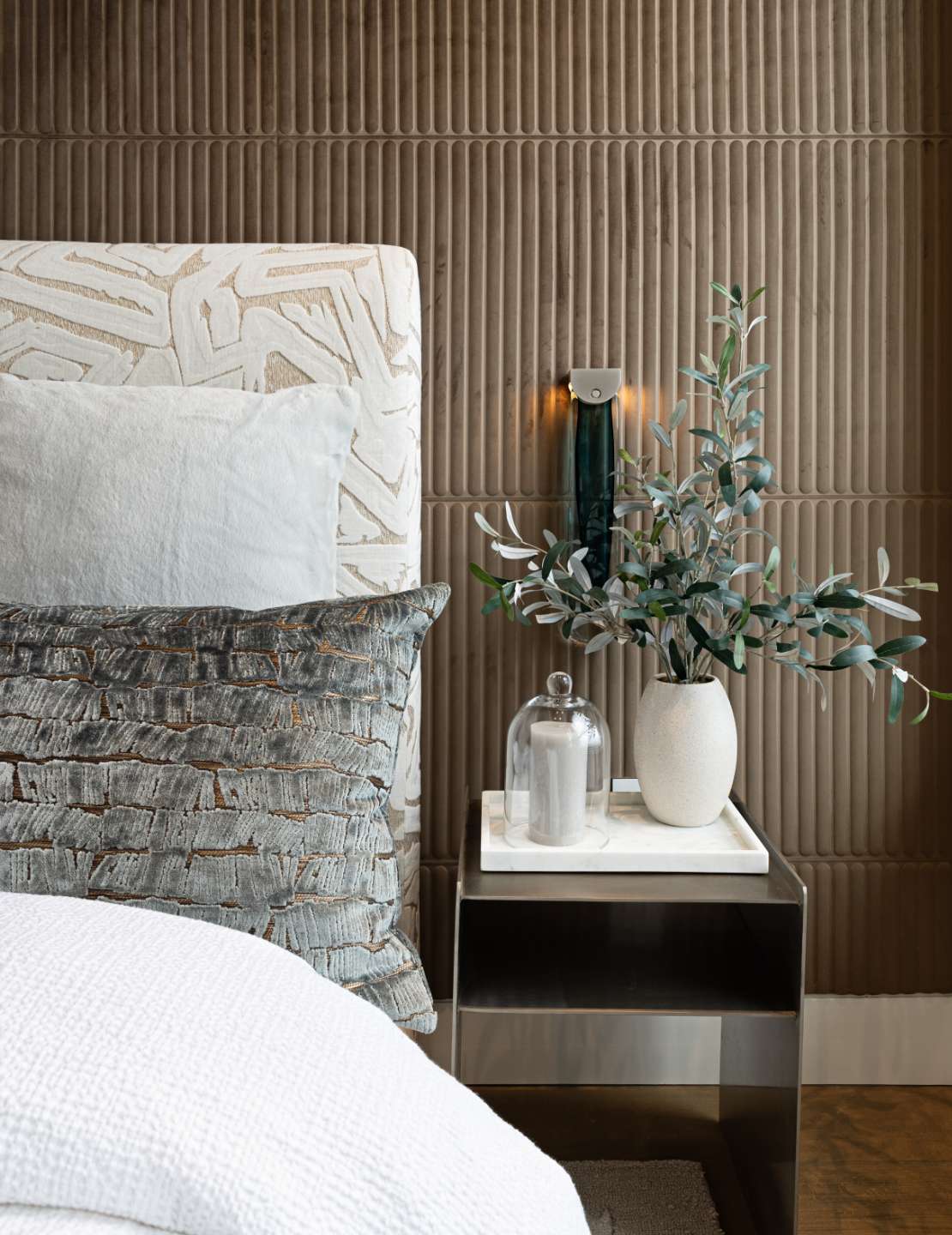 A bed and metal nightstand are shown against a wall with brown concrete treatment. A white vase and candle sit atop a marble tray on the nightstand.