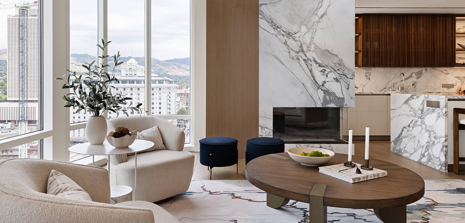 A living room with large windows is decorated with eggshell fabric lounge chairs and a modernist coffee table. A fireplace and kitchen area are shown with marble accents.