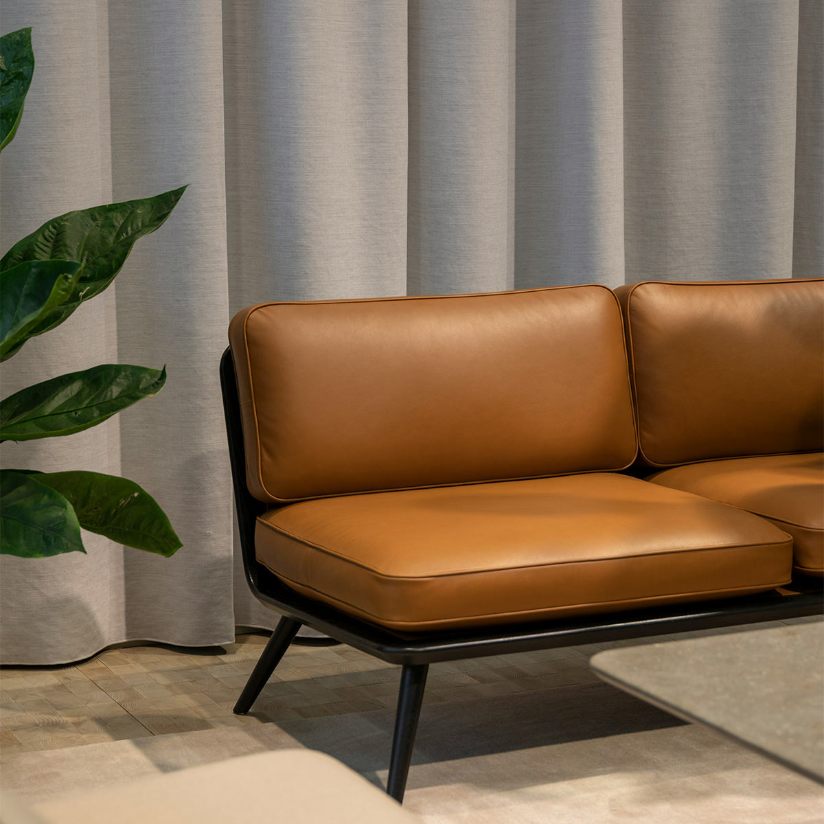 The Spine Lounge Suite Sofa designed by Space Copenhagen is shown with a black lacquer wood finish and caramel leather against a gray linen curtain.