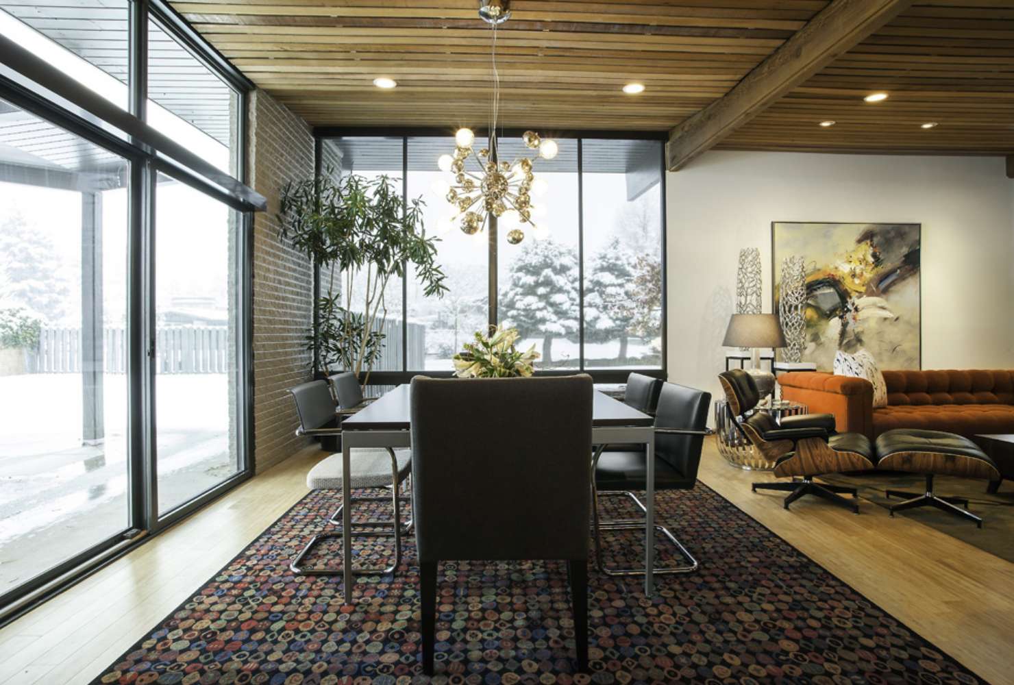 An open-concept dining room and adjoining living room are decorated with midcentury modern furniture, fine art, and an exposed wood beam ceiling.