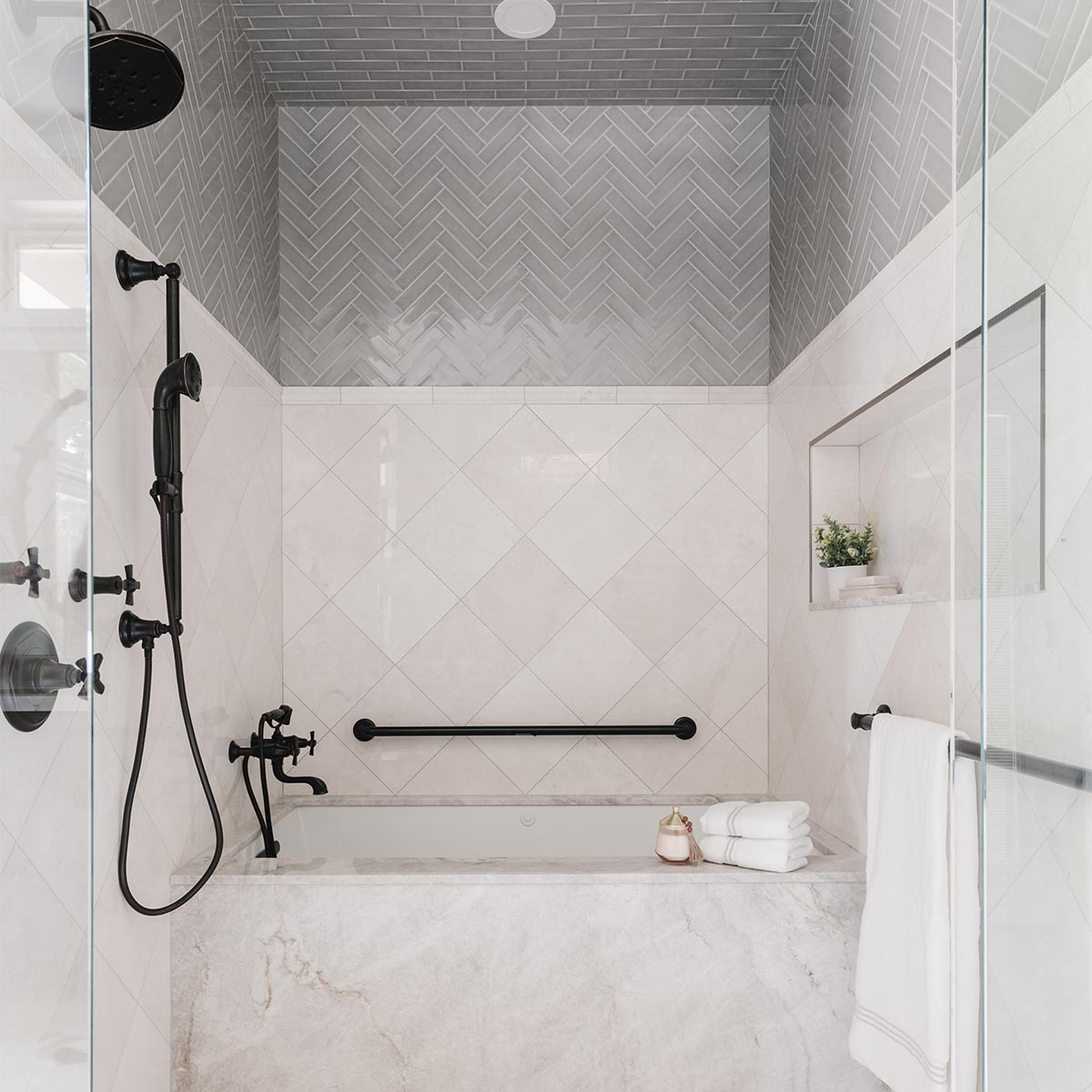 A shower and bathroom is shown with lightly pink marble coverings and cross-hatched gray subway tile. Dark fittings adorn the space to create a pleasing contrast. 