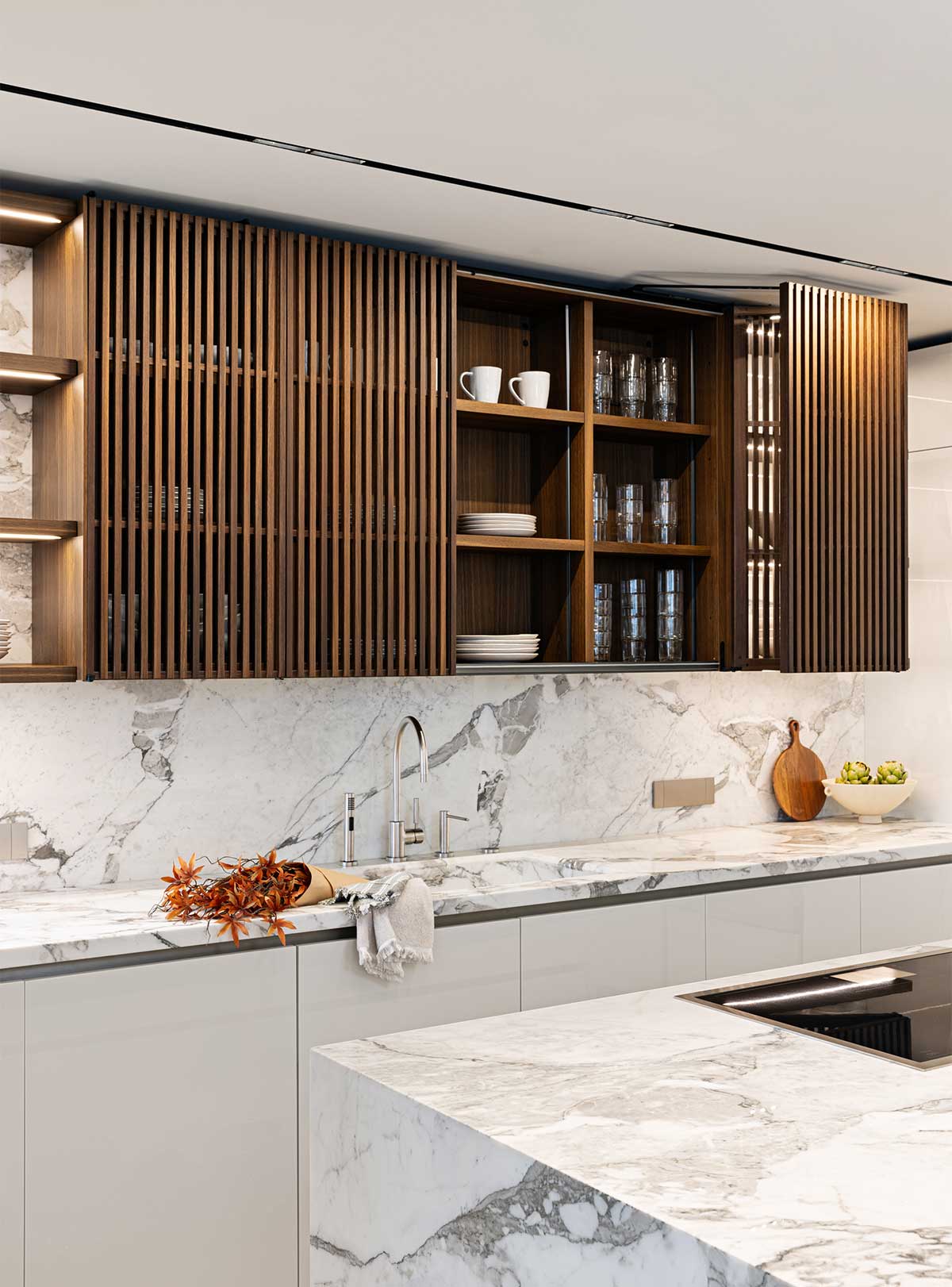 A kitchen space is shown with marble countertops and polished steel fittings. Oak-style wood cabinetry is shown with vertical-slatted doors that swivel out.