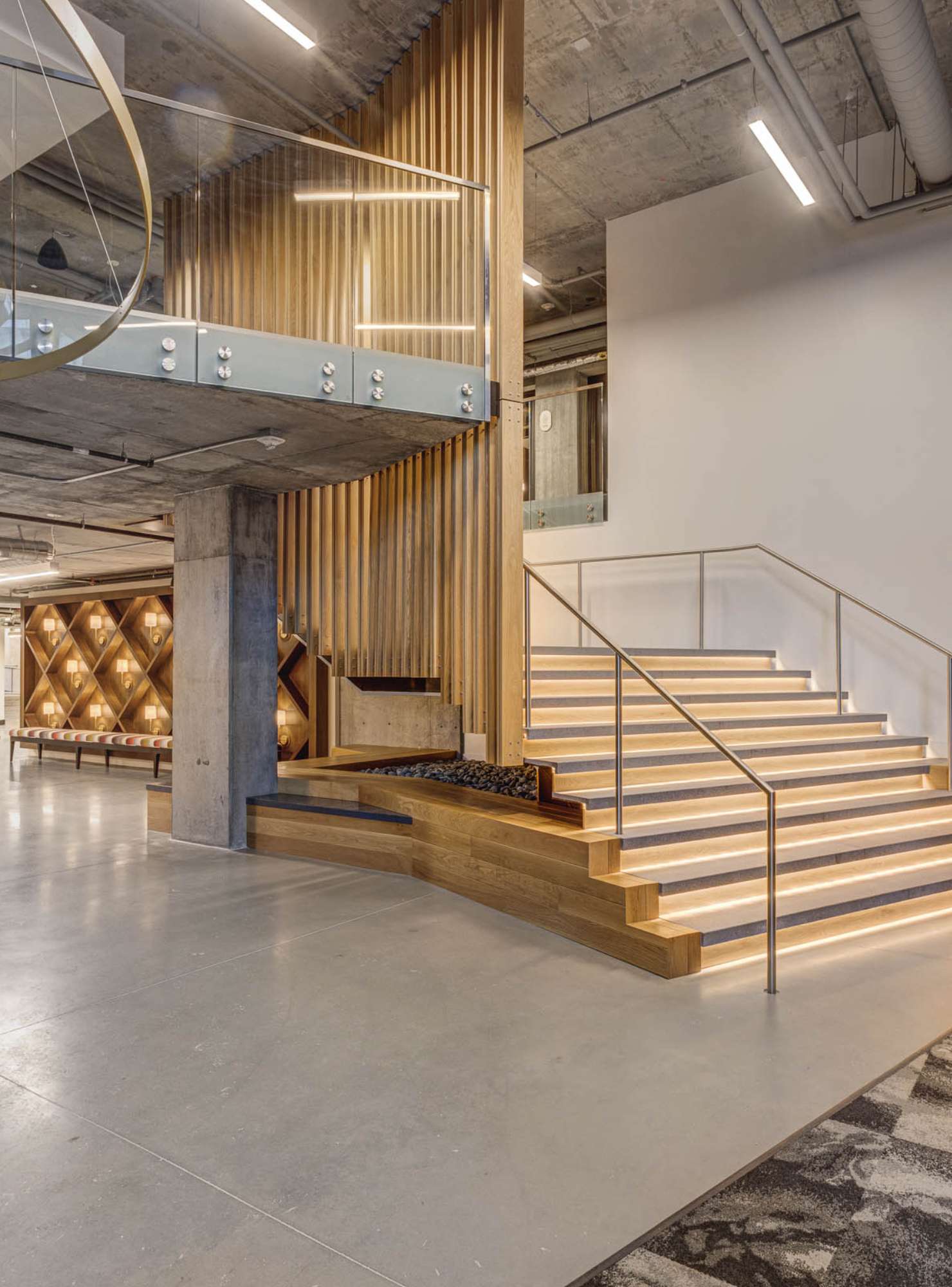A large office space is shown with a concrete pillar holding up an open balcony, a vertically slatted wood room divider, and a large staircase with built-in accent lighting.