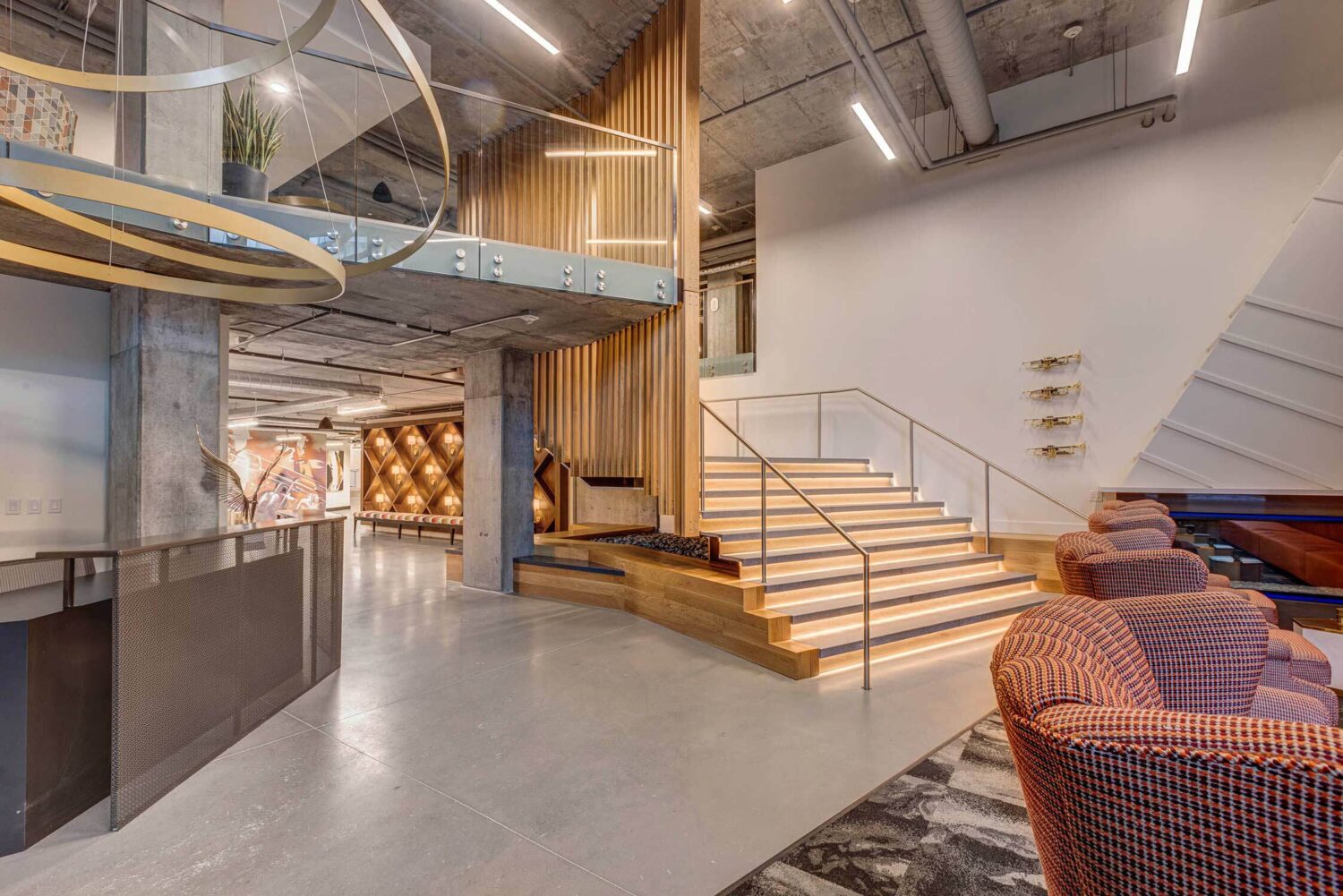 A modern staircase with illuminated steps ascends beside a wooden slatted wall in a spacious lobby, featuring a large spiral light fixture and contemporary chairs.