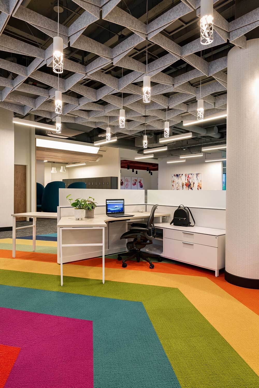 A modern desk sits with a computer displaying a cityscape wallpaper beside a potted plant and bag, surrounded by colorful geometric carpet and contemporary office decor.
