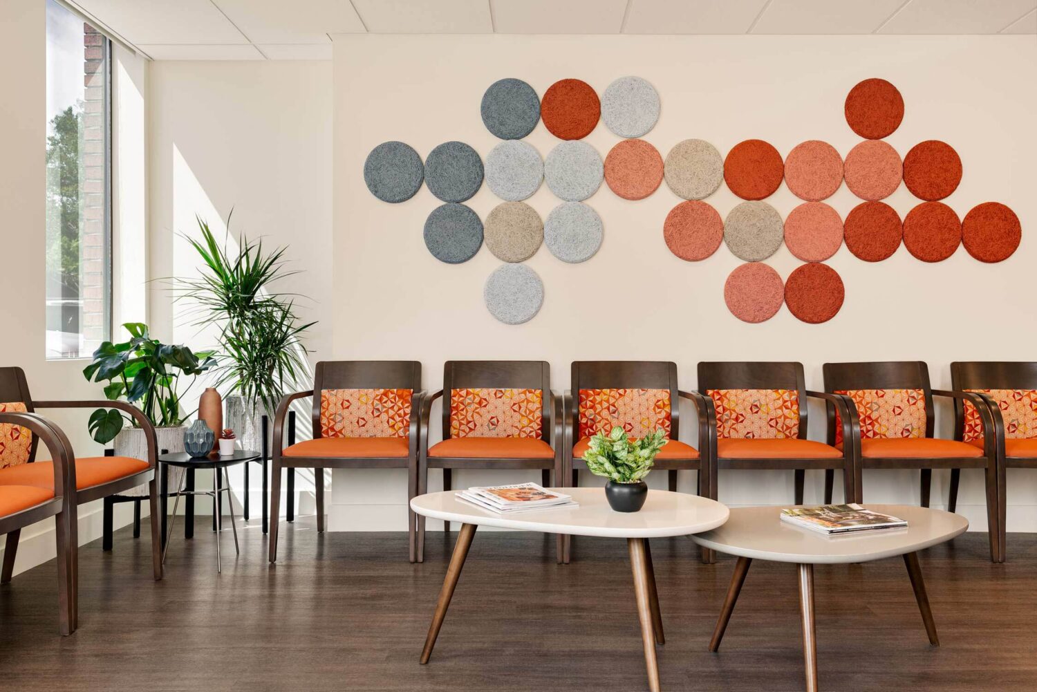 Chairs arranged in a waiting area feature patterned cushions. Circular wall decorations in various colors hang above. A potted plant and round tables with magazines enhance the room's modern decor.