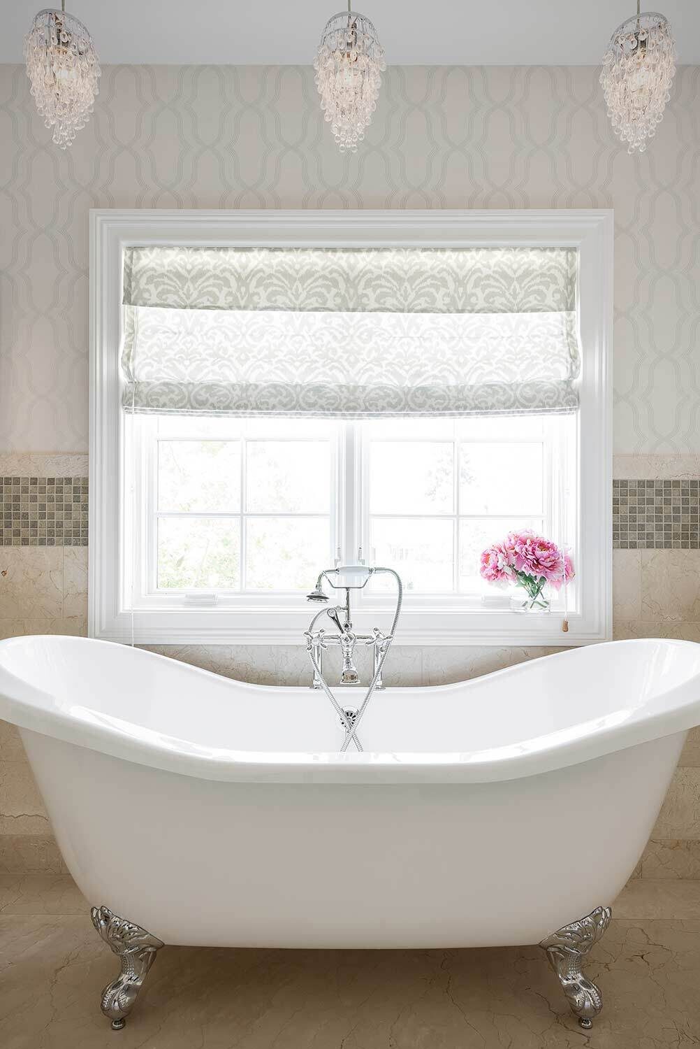 A white freestanding bathtub sits under a large window with floral curtains, surrounded by elegant cream tiles. Pink flowers in a vase decorate the windowsill, enhancing the bathroom's brightness.