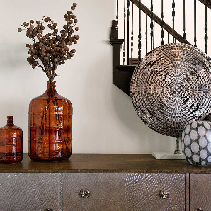 Vases with dried branches and a woven basket rest on a wooden sideboard near a staircase, creating a rustic decorative setting.