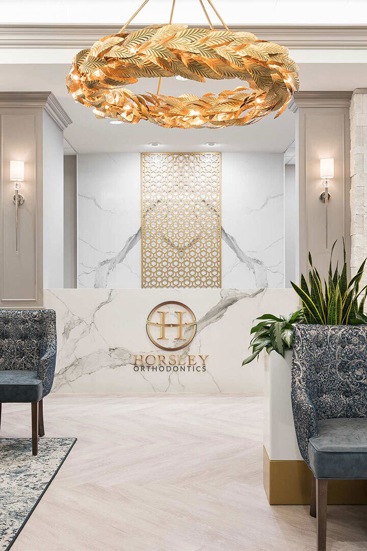 A large, decorative gold chandelier hangs above a modern reception area. Marble walls and a counter feature the logo "Horsley Orthodontics." Stylish furniture and plants enhance the elegant design.