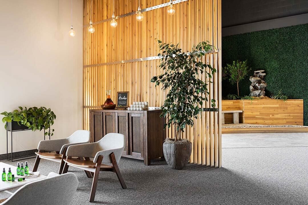 Two gray lounge chairs face a small table with green bottles in a modern office space. A wooden divider with hanging lights and a plant separates the room, featuring decorative elements.