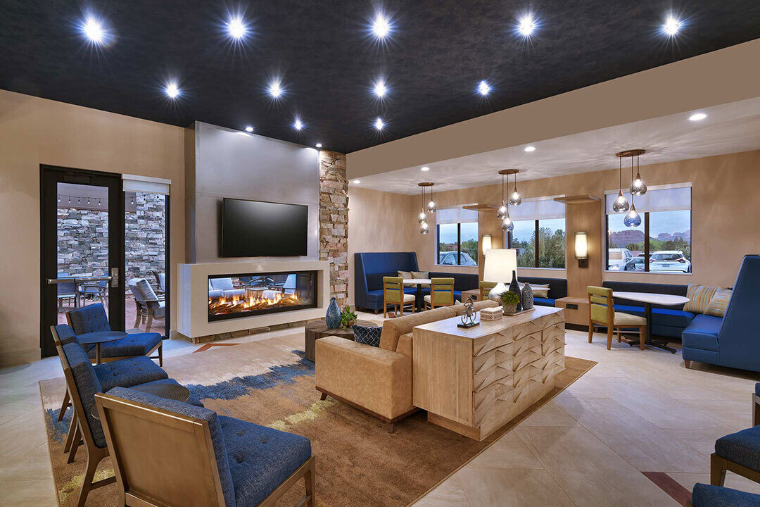 A modern lounge with blue chairs and beige sofas surrounds a central coffee table. A wall-mounted TV above a fireplace adds focus, while large windows provide natural light.