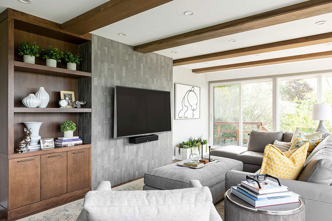 A living room decorated with a television, art, and plants. There is a grey sectional couch with an adjacent ottoman. Next to the televeision there is a cabinet with various vases and books. There is a glass door leading outisde.