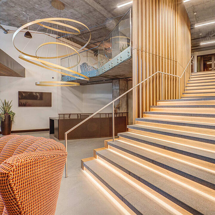 A modern staircase with illuminated steps ascends beside a wooden slatted wall in a spacious lobby, featuring a large spiral light fixture and contemporary chairs.