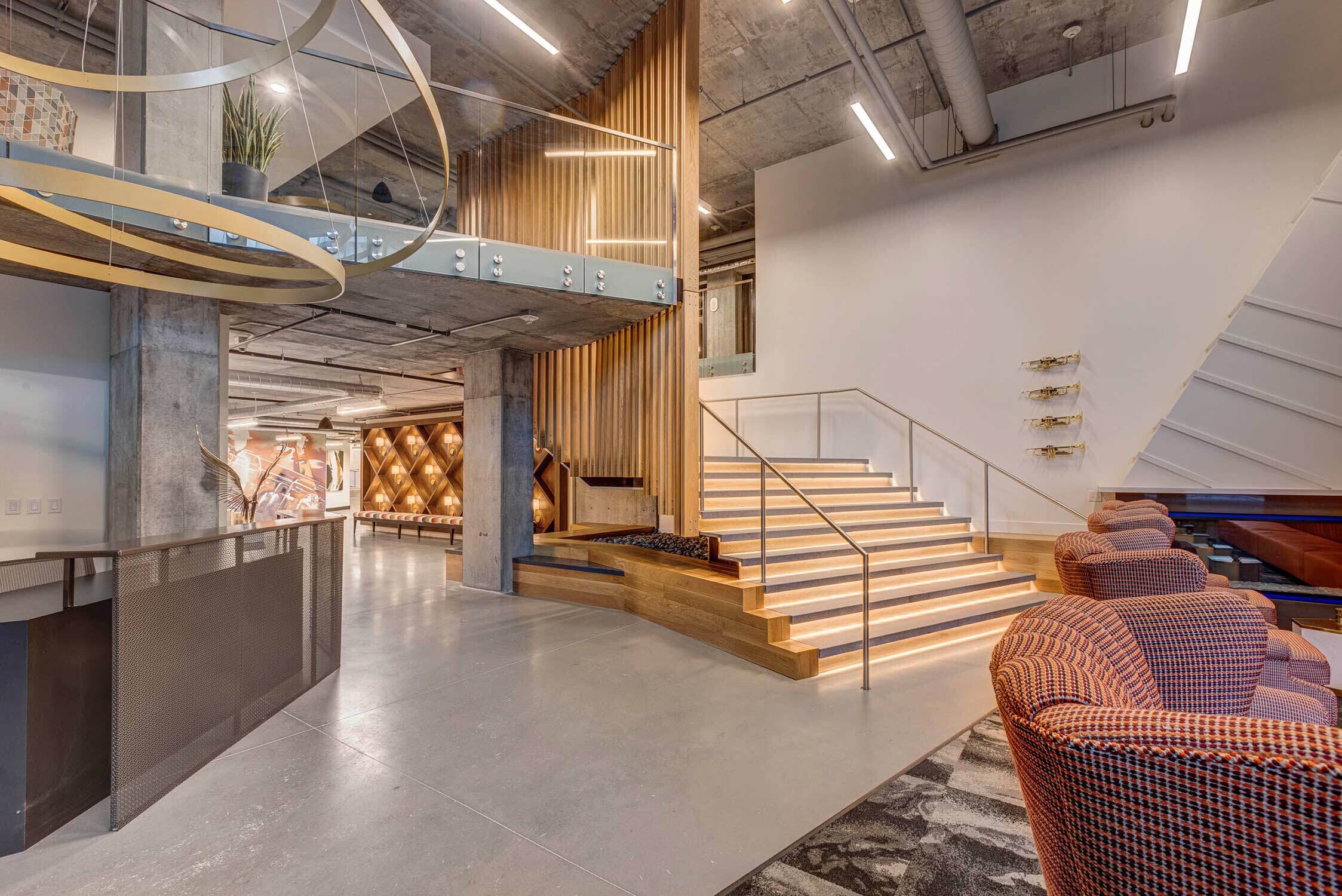 A modern staircase with illuminated steps ascends beside a wooden slatted wall in a spacious lobby, featuring a large spiral light fixture and contemporary chairs.