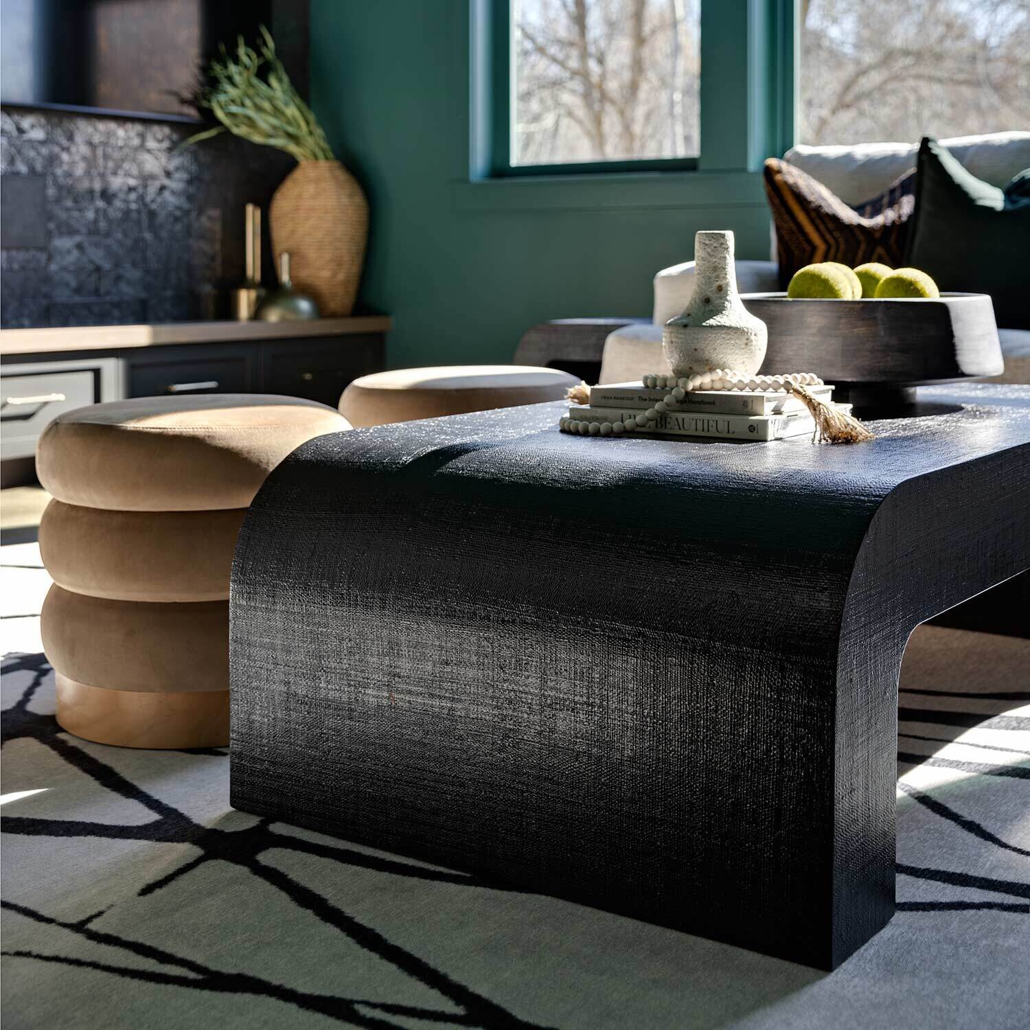 A modern black coffee table holds decorative items, including a vase and tray, in a stylish, sunlit living room with green walls, patterned rug, and large windows.
