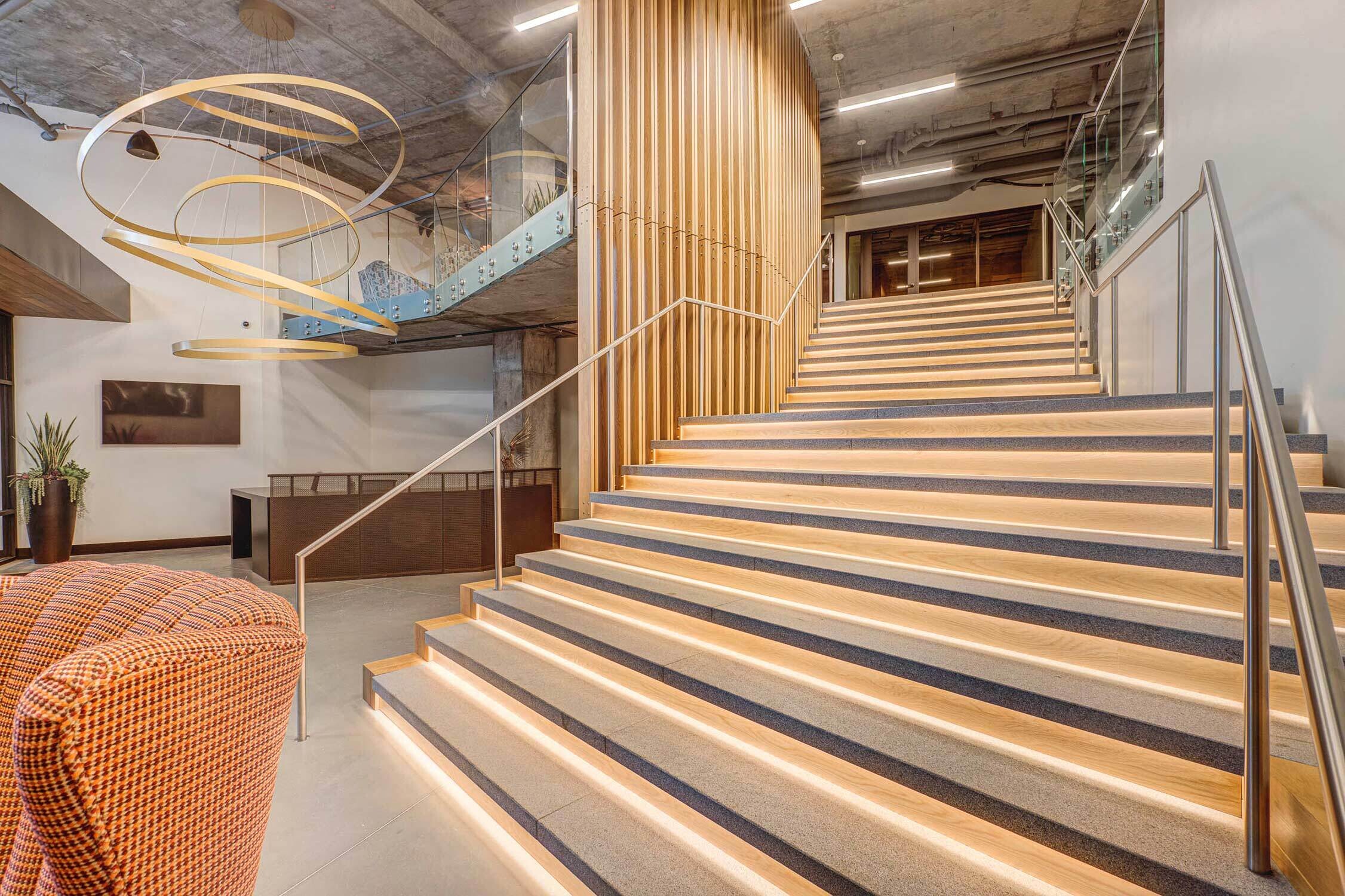 A modern staircase with illuminated steps ascends beside a wooden slatted wall in a spacious lobby, featuring a large spiral light fixture and contemporary chairs.