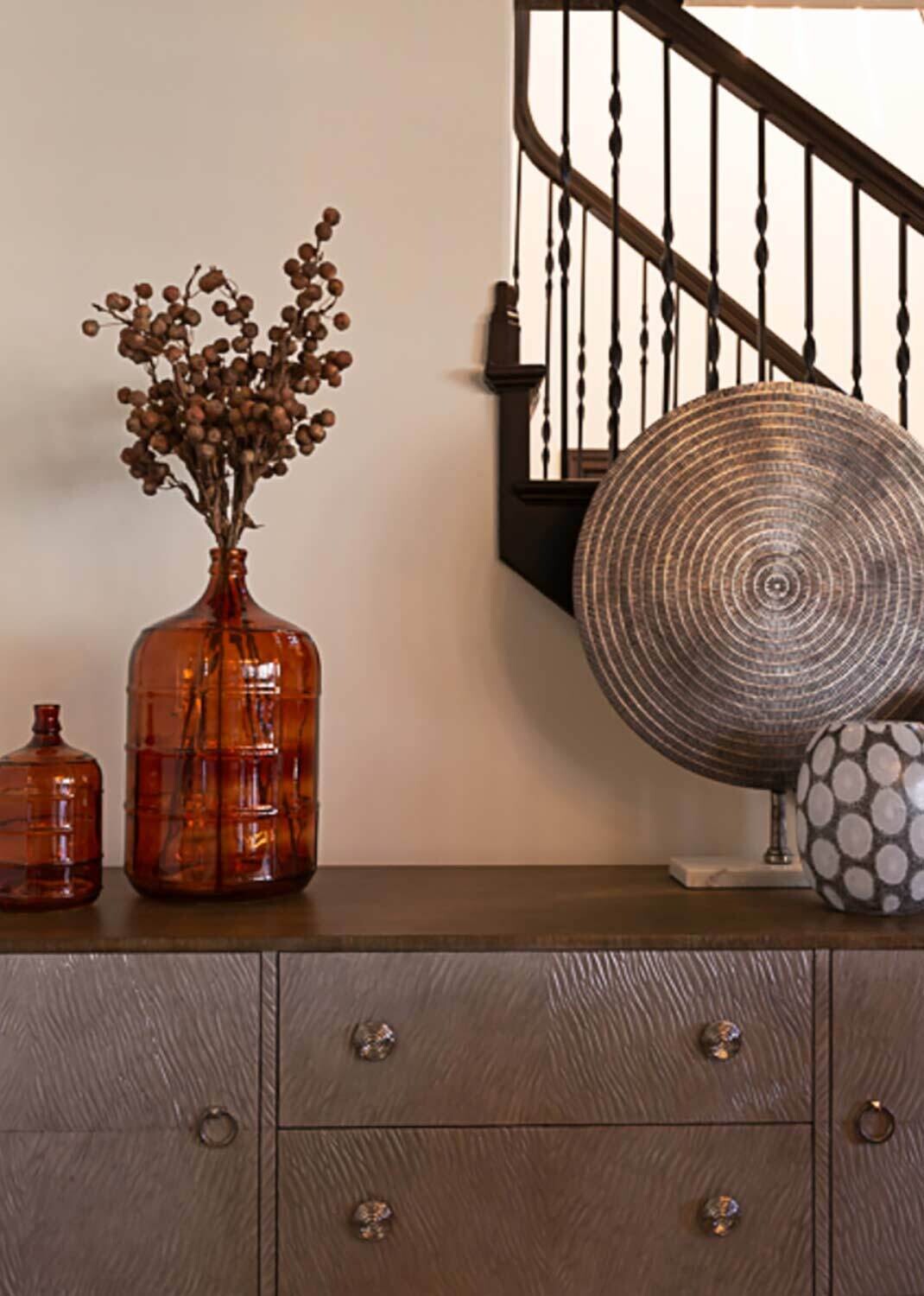 Vases with dried branches and a woven basket rest on a wooden sideboard near a staircase, creating a rustic decorative setting.