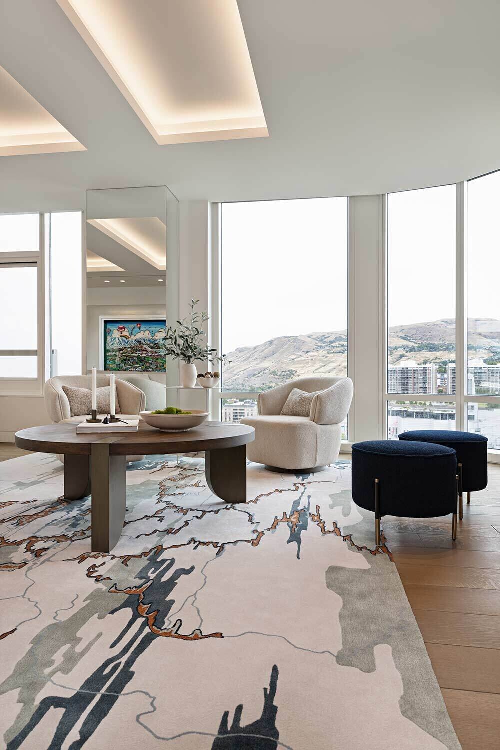 A living room overlooking the mountains decorated with two cream arm chairs, two blue ottoman's, and a dark brown coffee table. The coffee table is decorated eith candles and books. There is also a small side table between the chairs with decorative plants on it.