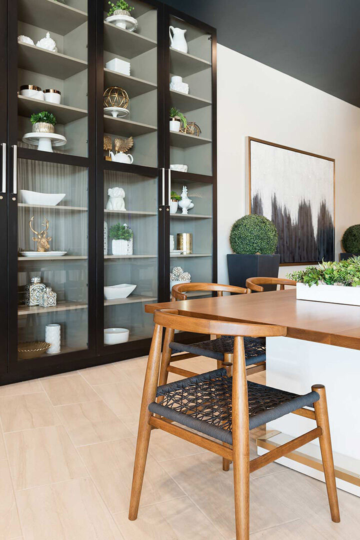 A wooden dining table with woven chairs is set near glass cabinets displaying decorative items, like vases and plants, in a modern room with light flooring and artwork on the wall.