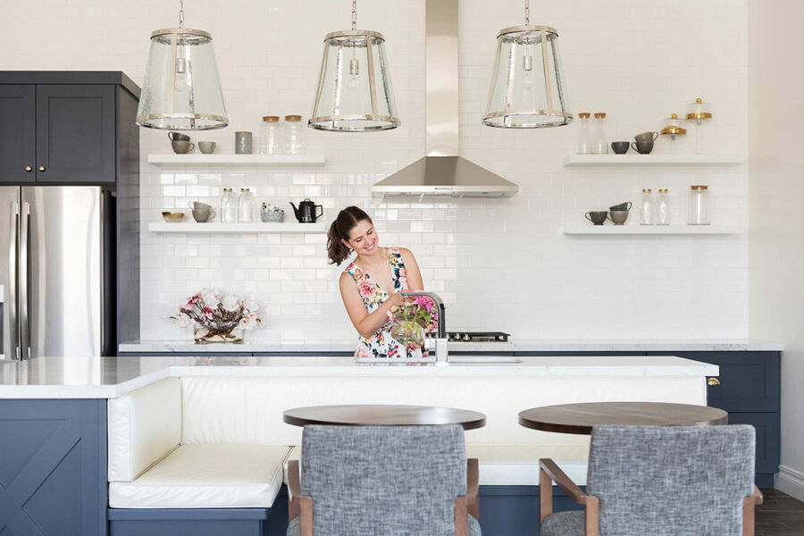 A woman arranges flowers in a vase on a kitchen island with white countertops. The kitchen features dark cabinets, stainless steel appliances, open shelves, and pendant lights, creating a modern atmosphere.