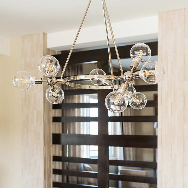 A modern chandelier with exposed bulbs hangs from the ceiling, centered within a contemporary living space featuring neutral walls and dark wooden accents in the background.