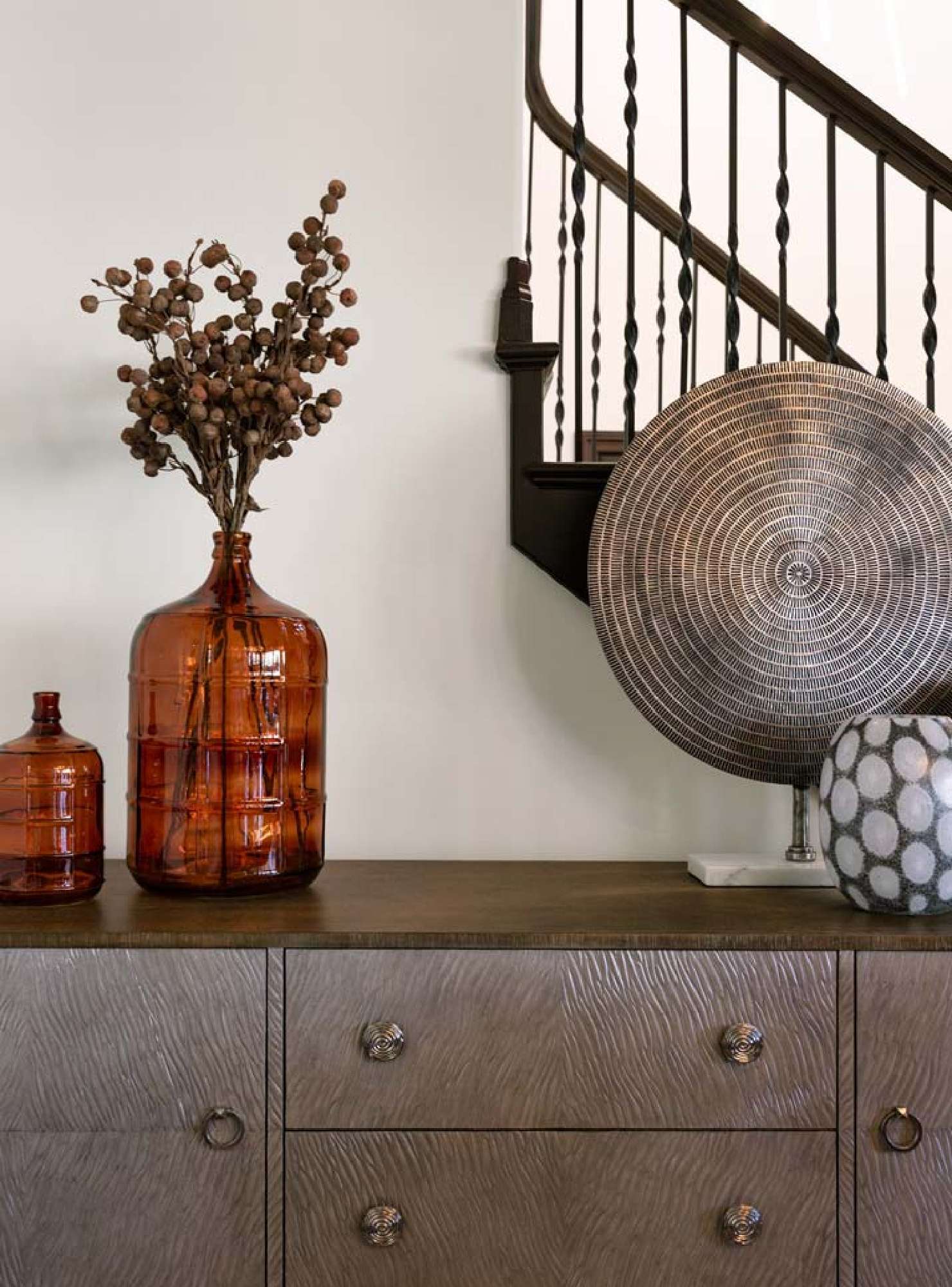 Dark glass vases and a circular brasswork sculpture sit atop a wood and brass entryway credenza